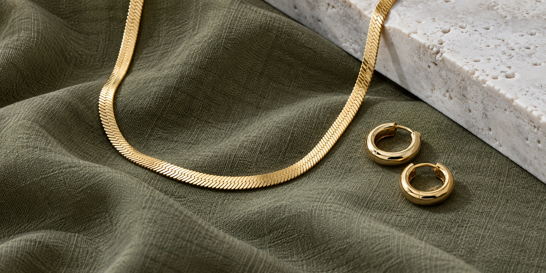 Gold herringbone necklace and hoop earrings arranged on muted olive fabric