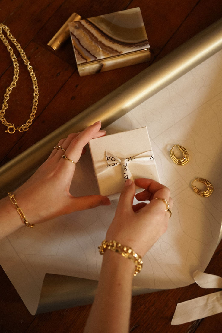 Hands wrapping a gift box beside gold hoop earrings, a chain necklace and bracelets