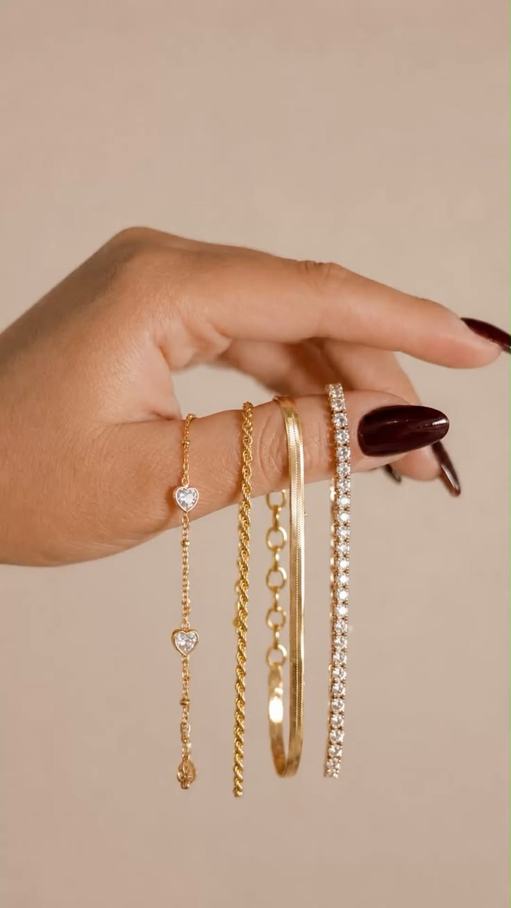 Hand with burgundy nails holding a gold heart station bracelet, a rope chain, a herringbone bracelet and a diamond tennis bracelet