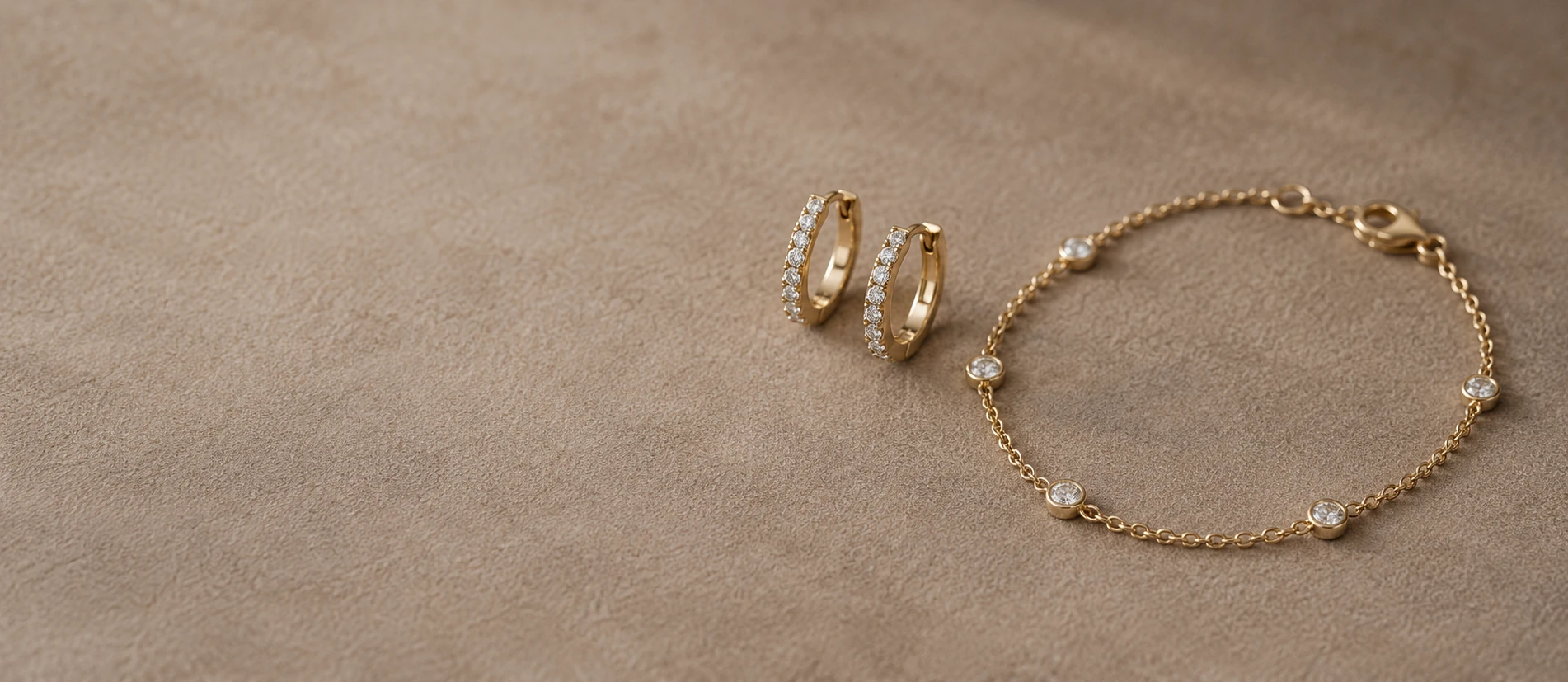 Yellow-gold diamond hoop earrings and a delicate diamond station bracelet on warm taupe suede in soft daylight