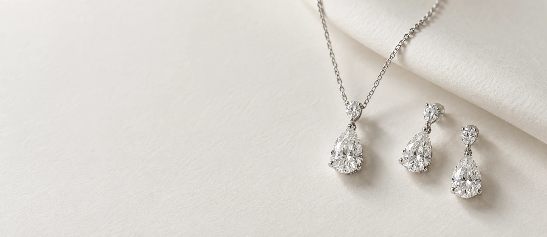 White-gold pear-cut diamond pendant necklace and matching drop earrings on softly folded ivory paper in natural daylight